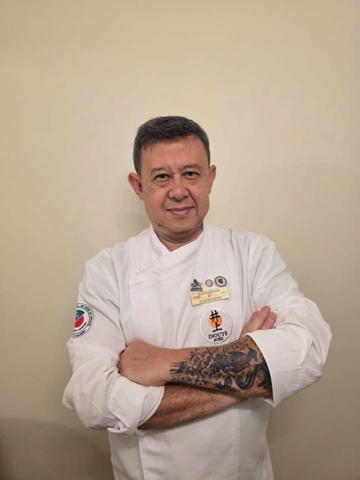 Chef Inouye de dólmã branco, braços cruzados, com crachás de certificação e o patch Inouye Sushi